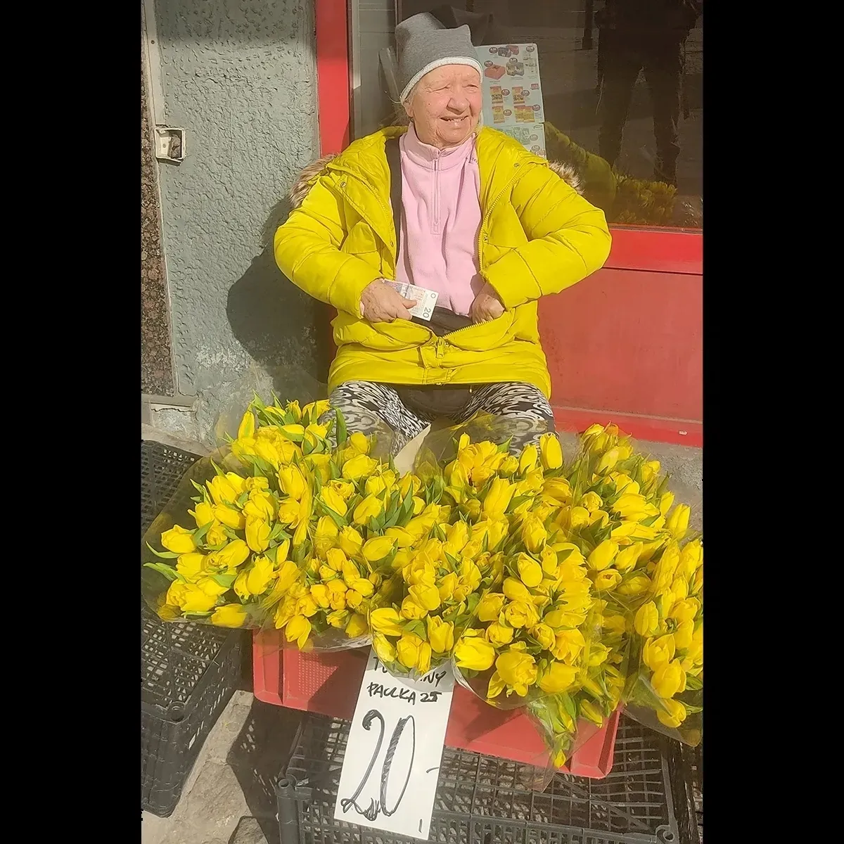Older flower seller in a yellow jacket sitting behind bunches of yellow tulips